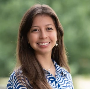 This image depicts a young women smiling at the camera. She is in front of trees. 